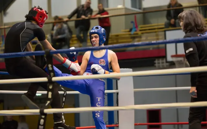 Un boxeur du TCMS en tenue bleue lève la jambe pour un coup de pied, sur le ring, face à son adversaire.